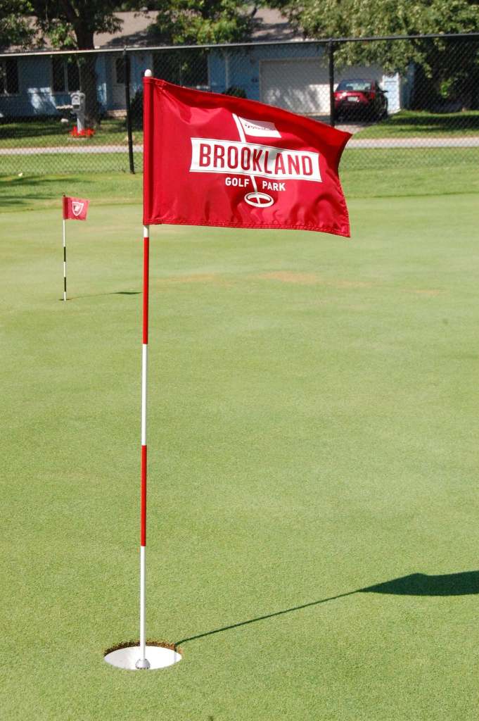 A red Brookland Golf Park flag waving in the wind planted in the hole for a green.